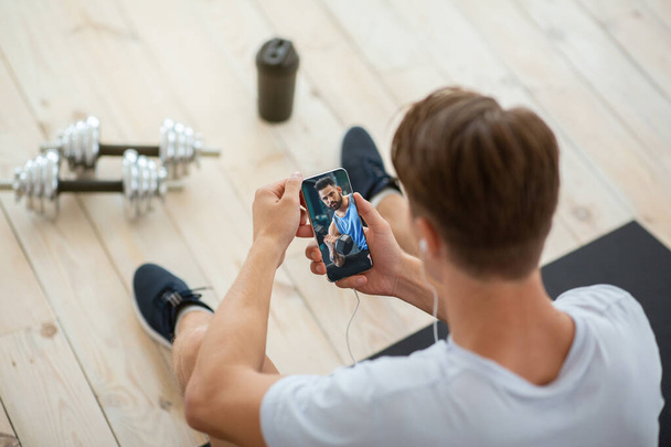 Online προπονητής για προπόνηση στο σπίτι. Αγνώριστος νεαρός που παρακολουθεί μαθήματα βίντεο bodybuilding στο smartphone - Φωτογραφία, εικόνα