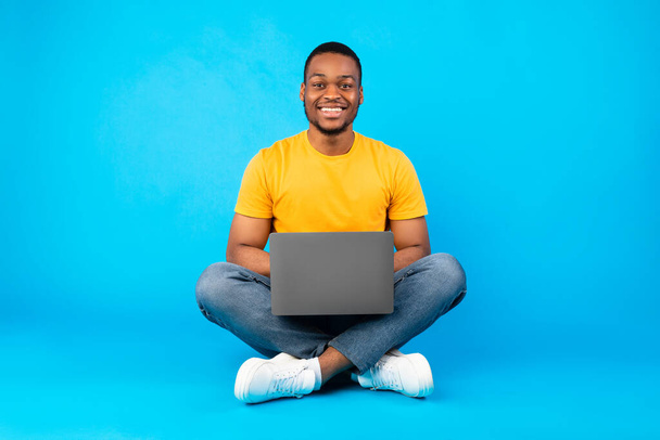 feliz afro-americano cara sentado com laptop sobre fundo azul - Foto, Imagem
