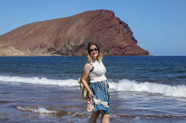 Retrato de una joven milenaria exuberante con un vestido de paisley azul y blanco mientras sonríe, camina y disfruta de sus días de vacaciones de verano en la playa de La Tejita, Tenerife, Islas Canarias, España - Foto, Imagen