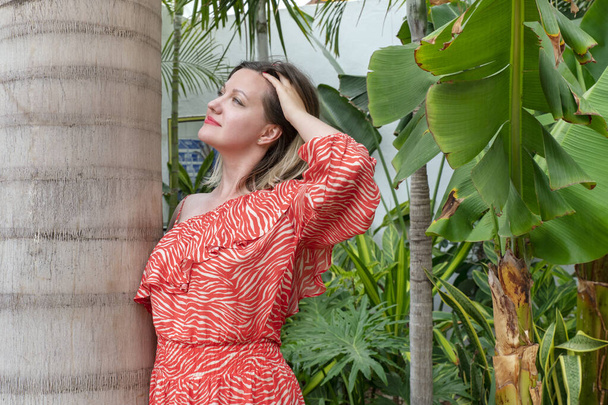 Ritratto sognante di una giovane donna attraente millenaria che indossa un abito rosso estivo mentre si appoggia a una palma e passa le dita tra i capelli, godendo e rilassandosi in un giardino tropicale - Foto, immagini