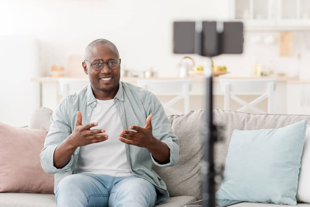 Vrolijke volwassen Afrikaans-Amerikaanse man uitzenden vanuit huis, met behulp van smartphone en statief, zitten op de bank thuis - Foto, afbeelding