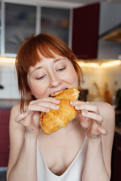 Κορίτσι τρώει κρουασάν με όρεξη, ενώ το πρωινό στο φως της κουζίνας - Φωτογραφία, εικόνα