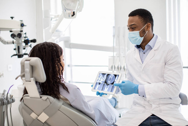 Male Black Dentist Doctor Demonstrating Female Patient Teeth Xray On Digital Tablet - 写真・画像