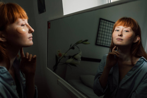 Female is looking into the mirror while wearing eye patches against wrinkles - 写真・画像