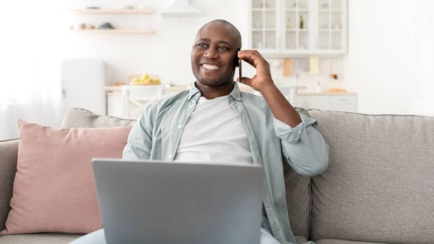 Mobile communication. Smaling african american man using laptop and talking on cellphone, sitting on couch - Фото, изображение