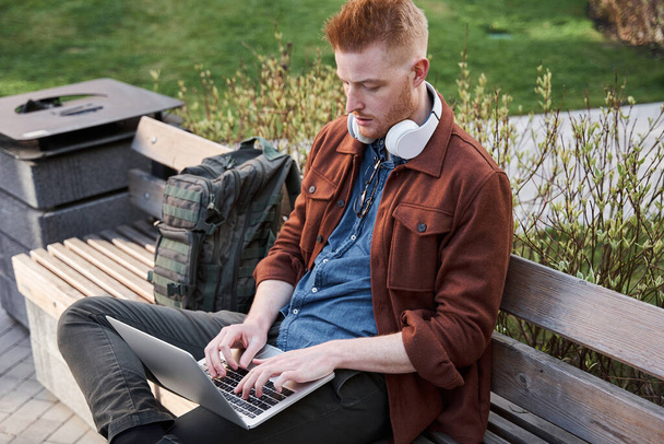 Hipster man with laptop sitting on the bench at the street and working remotely - Photo, Image