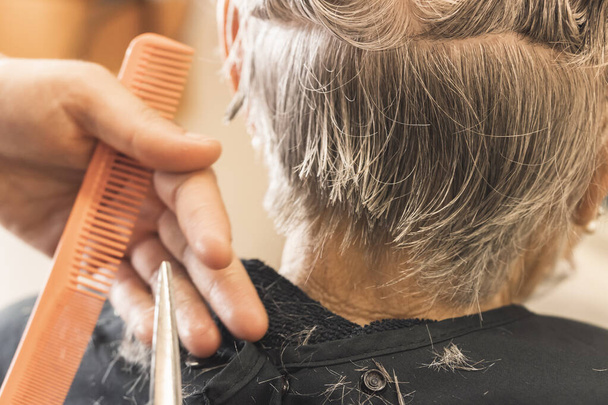 Крупный план женщины-стилиста, обрезающей ножницами и оранжевой расчёской влажные волосы пожилого клиента в парикмахерской на размытом фоне. - Фото, изображение