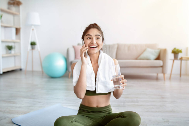 Sportos fiatal indiai hölgy egy pohár vízzel beszél a mobilon, miután szünetelteti az otthoni edzés - Fotó, kép
