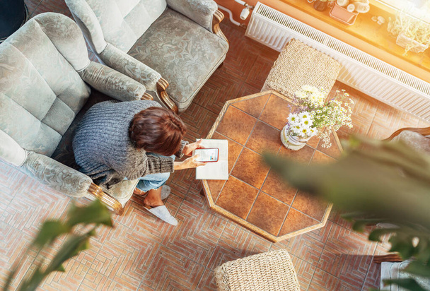 Top view of a female dressed cozy home clothes in comfortable armchair, browsing an internet using smartphone in house sunroom living room. Stay-at-home or business home office concept image - 写真・画像