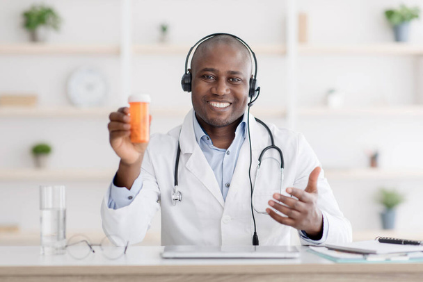 Doctor recommendation. Happy african american therapist in white coat showing jar of pills, having online consultation - Фото, зображення