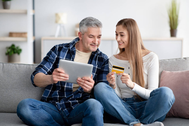 Online shopping concept. Cheerful mature couple using digital tablet and credit card together at home - Photo, image