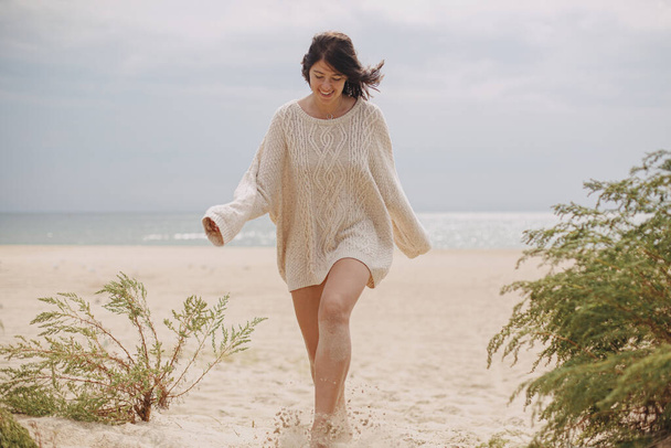 Счастливая женщина с ветреными волосами, гуляющая по песчаному пляжу на фоне зеленой травы и моря, спокойный спокойный момент. Стильная молодая женщина в бежевом трикотажном свитере отдыхает на холодном побережье - Фото, изображение