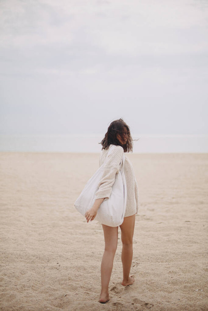 Schöne stilvolle Frau mit windigen Haaren und Tragetasche, die am Sandstrand bis zum Meer spaziert, unbeschwerter Moment. Stilvolle junge Frau im Strickpullover genießt Urlaub und Entspannung an der Küste. Vertikales Bild - Foto, Bild