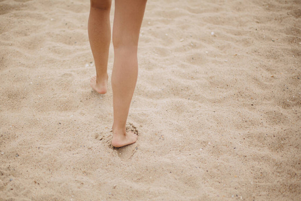 Hermosas piernas de mujer descalza caminando en la playa de arena, estado de ánimo de vacaciones sin preocupaciones. Elegante joven hembra relajante en la costa, vista recortada. Playa de verano. Espacio para texto - Foto, Imagen