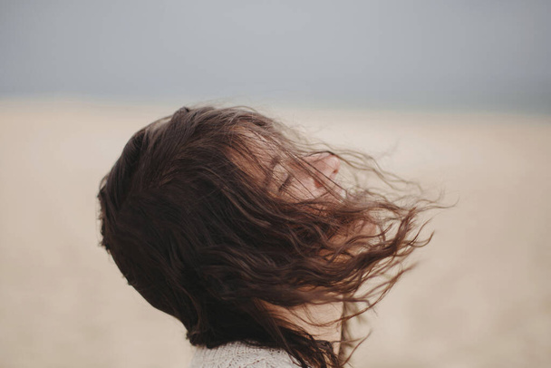 Carefree beautiful woman with windy hair on background of sandy beach and sea, tranquil moment. Portrait of stylish young female relaxing and enjoying vacation on coast - Foto, imagen