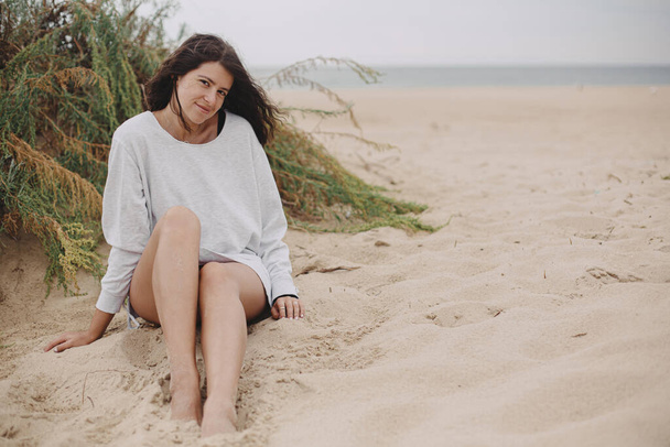 風の強い髪を持つ美しい女性とセーターの緑の芝生と海、穏やかな静かな瞬間を背景に砂浜に座っている。海岸でリラックスしたスタイリッシュな若い女性。休暇気分 - 写真・画像