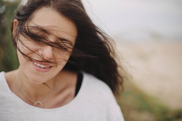 Beautiful happy woman with windy hair portrait on background of sandy beach and grass, carefree tranquil moment. Stylish young female in sweater relaxing and enjoying vacation on coast - Photo, Image