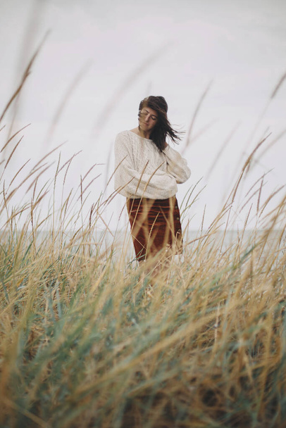 Όμορφη κομψή γυναίκα σε πλεκτό πουλόβερ και με θυελλώδη μαλλιά ποζάρουν ανάμεσα σε άγριο χορτάρι στη θάλασσα. Αυθεντική ανέμελη στιγμή. Μοντέρνο νεαρό θηλυκό που στέκεται στην ανεμοδαρμένη ακτή. Ηρεμία. Κάθετη - Φωτογραφία, εικόνα