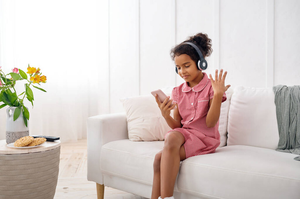 маленькая девочка в наушниках слушает музыку дома. девочка сидит на диване и с помощью мобильного телефона, смешной ребенок с современным гаджетом - Фото, изображение
