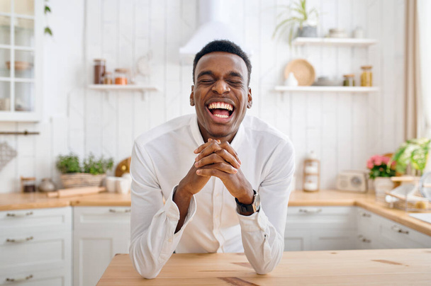 Laughing man sitting at the counter on the kitchen. Cheerful male person poses at the table at home in the morning, happy lifestyle - Foto, afbeelding