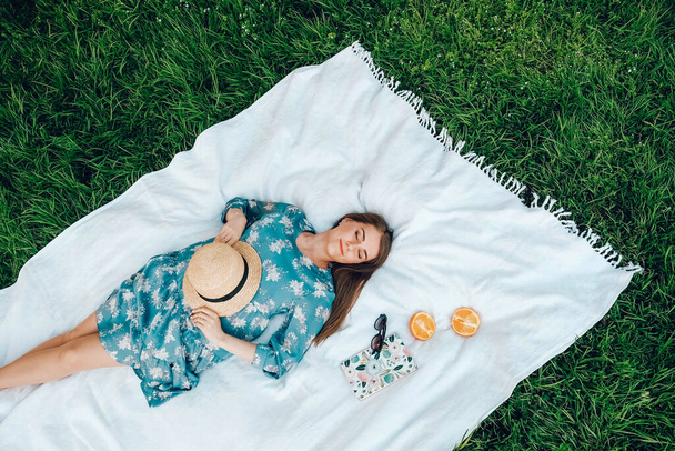 Renkli elbiseli ve hasır şapkalı güzel bir kadın yeşil çimen arka planında beyaz bir battaniyenin üzerinde uzanır, kadın aksesuarları, yaz zamanı. Kopyala, metin için boşluk. - Fotoğraf, Görsel