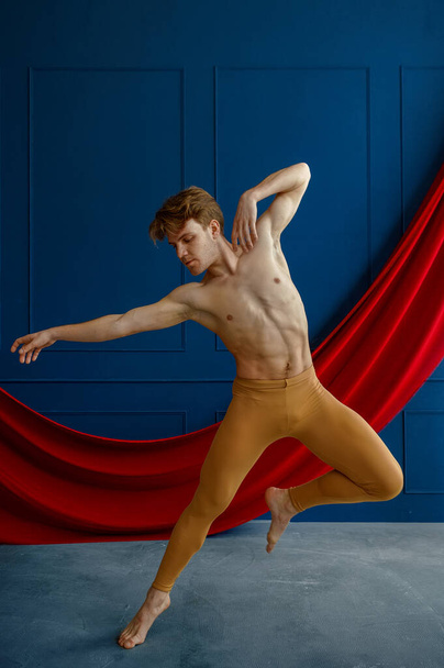 Mužský baletní tanečník, trénink ve třídě tance, modré stěny a červená látka na pozadí. Účinkující s svalnatým tělem, elegancí pohybů - Fotografie, Obrázek
