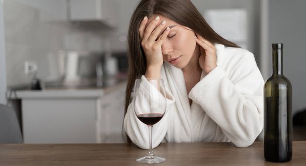 アルコール依存症。一人の女性が自宅のキッチンでワインを飲んでいると深く落ち込んでいる。アルコールの問題と解決策.少女や悲しみの孤独. - 写真・画像