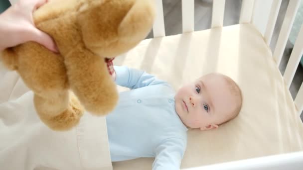 Mother giving plush teddy bear to her little baby boy lying in cradle. Concept of parenting, family happiness and baby development - Footage, Video