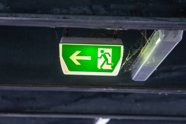 Zielony znak wyjścia z osobą biegnącą i zieloną strzałką pokazuje oznakowanie systemu naprowadzania na parkingu dla ratowania i ewakuacji bezpieczeństwa w niebezpiecznych sytuacjach na suficie ratuje życie w skrajnym niebezpieczeństwie - Zdjęcie, obraz