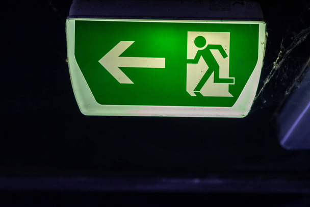 Groene exit bord met lopende persoon en groene pijl toont geleidingssysteem bewegwijzering op een parkeerplaats voor redding en evacuatie veiligheid in gevaarlijke situaties aan het plafond redt leven in extreem gevaar - Foto, afbeelding