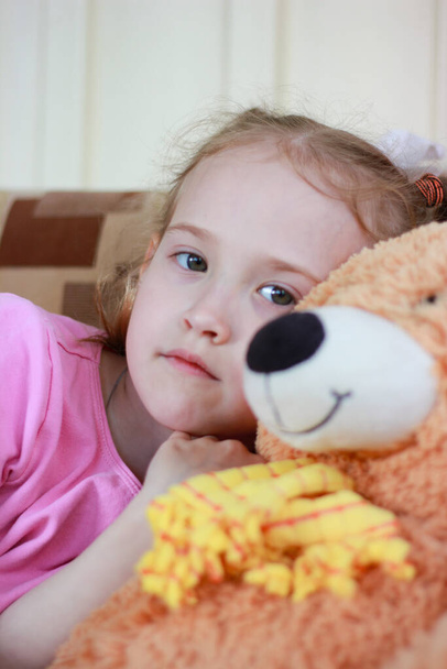 little girl with sad eyes looks at the camera sits with an orange bear toy - Foto, Bild