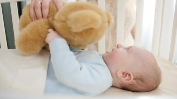 Happy smiling baby boy palying and holding teddy bear while lying in wooden cradle. Concept of parenting, family happiness and baby development - Footage, Video