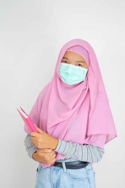 Estudiante joven usar rosa hijab y máscara celebrar libros sobre fondo blanco. - Foto, imagen