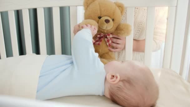 Toy teddy bear playing with little baby boy lying in cradle. Concept of parenting, family happiness and baby development - Footage, Video