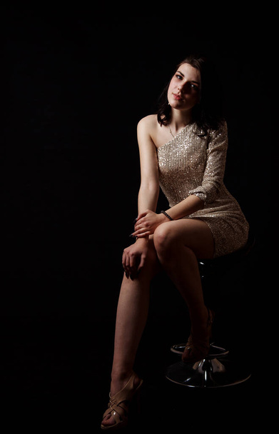 joven hermosa chica morena en vestido brillante posando sentado en la silla en el estudio sobre fondo negro - Foto, imagen