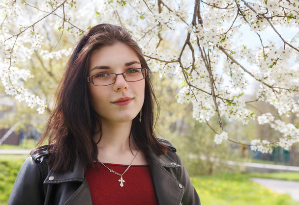 portrait de jeune belle fille brune en veste noire et pull bordeaux debout près des fleurs de cerisier le jour ensoleillé du printemps. gros plan extérieur - Photo, image