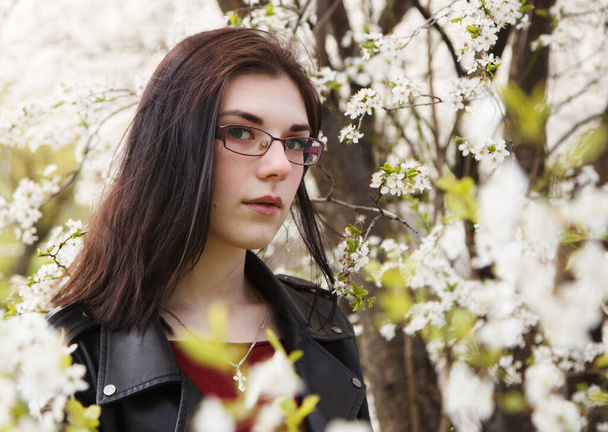 春の晴れた日に桜の近くに立って黒のジャケットとバーガンディセーターで若い美しいブルネットの女の子の肖像画。屋外の閉鎖 - 写真・画像