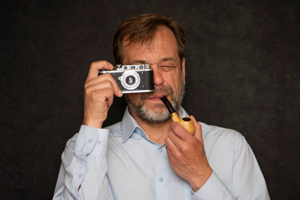 Retrato de un anciano de 45-50 años con una pipa humeante en la boca y una camisa clara sobre un fondo oscuro, tomando fotos con una cámara vieja. - Foto, imagen