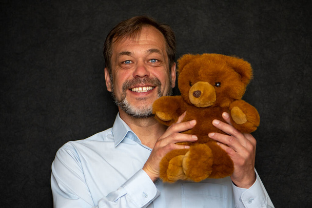 Un homme souriant âgé de 45 à 50 ans avec une barbe dans une chemise légère tient un ours en peluche sur un fond sombre. Concept : complexes pour adultes, cadeaux pour enfants, - Photo, image