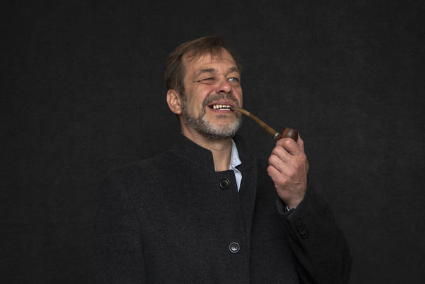 Portrait d'un homme âgé souriant âgé de 45 à 50 ans avec une barbe grise en manteau noir fumant une pipe sur un fond sombre.Concept : soldat, écrivain ou marin à la retraite. - Photo, image