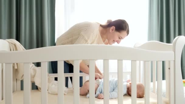 Krásná usměvavá matka svléká svého malého synka ležícího ráno v kolébce, aby vyměnila plenky. Pojem rodičovství, rodinné štěstí a vývoj dítěte. - Záběry, video