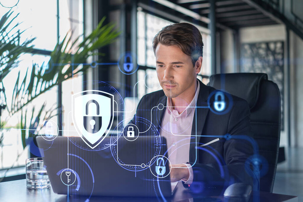 Handsome businessman in suit at workplace working with laptop to defend customer cyber security. Concept of clients information protection and brainstorm. Padlock hologram over office background. - Valokuva, kuva