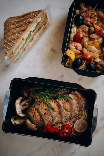 lunch delivery for two. duck with noodles and prawns on sticks with garnish. sandwich in a plastic container. fitness food in a paper container top view. - Фото, изображение