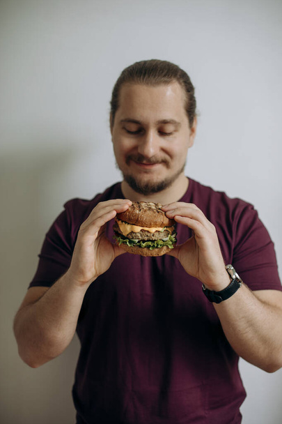 Een gelukkige man met een baard met een hamburger in zijn handen. De gelukkige gaat een heerlijke verse cheeseburger snack eten. De man kijkt verliefd op zijn hamburger voor hij eet. kopieerruimte. - Foto, afbeelding