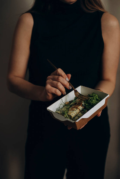 young woman holding food from delivery or catering in her hands. pancakes with spinach and herbs in a paper bowl. girl in dark clothes holds food from the delivery company. - Fotografie, Obrázek