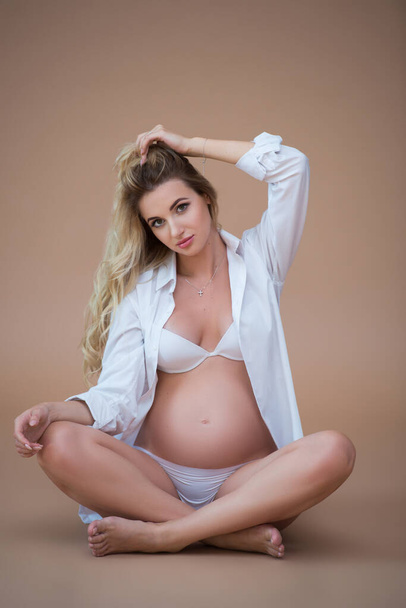 Beautiful pregnant blonde woman sits in a white shirt and underwear on a beige background and hugs her tummy with her hands. Healthy pregnancy. - Fotografie, Obrázek