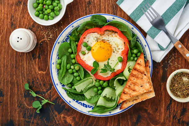 Red bell peppers stuffed with eggs, spinach leaves, green peas and microgreens on a breakfast plate on old wooden table background. Top view. - 写真・画像
