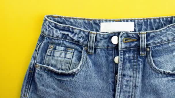 Bewegende foto van blauwe unisex jeans met metalen knopen, ritssluiting, zakken, wit label plat op gele achtergrond, kopieerruimte. Dagelijkse mode kleding in minimalistische stijl. Een casual outfit. Denim jeans - Video