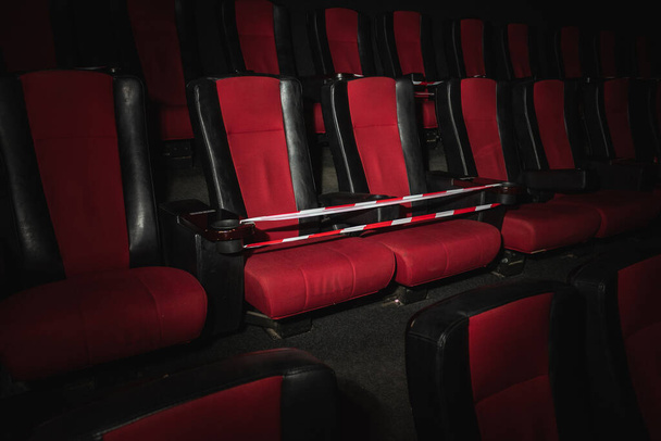 Empty movie theater. Armchairs red and black in the cinema hall are illuminated by a beam of light, low key. Selective focus. Quarantine, restrictive tape on chairs - Fotoğraf, Görsel