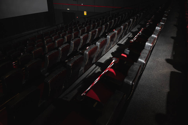 Cine vacío. Sillones rojos y negros en la sala de cine están iluminados por un haz de luz, bajo llave. Enfoque selectivo. Cuarentena, cinta restrictiva en sillas - Foto, imagen